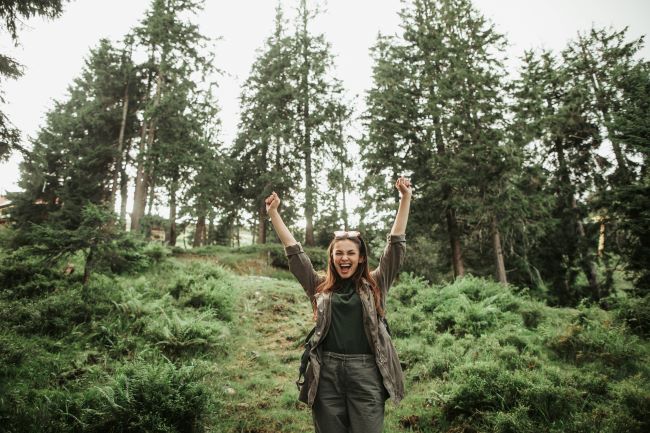 Mladá žena stojí na kopci v lese, ruce má zvednuté vzhůru jako náznak svobody.